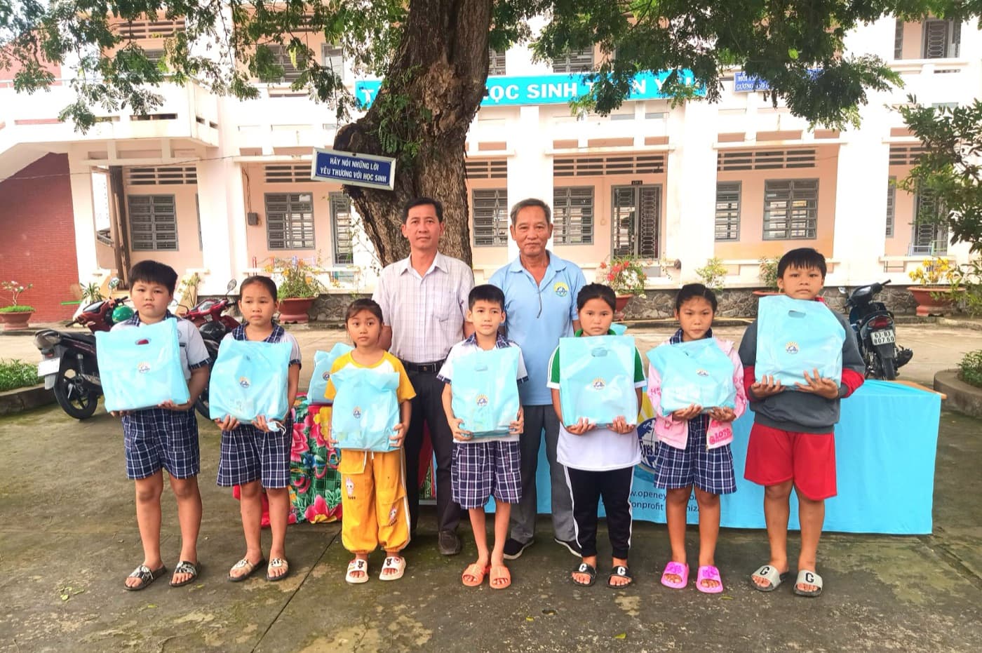 Schoolchildren in Vietnam holding OEBB school-support gift bags with an OEBB volunteer and their teacher